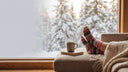 Pieds en chaussettes nordiques Polar Star sur un banc de fenêtre en bois, devant une baie ouverte sur une forêt enneigée, mug et livre