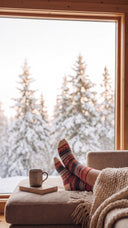 Pieds en chaussettes nordiques Polar Star sur un banc de fenêtre, grande baie ouverte sur des sapins enneigés, mug et livre